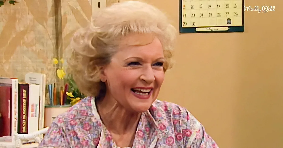 A smiling woman with blonde hair in a floral shirt sits in a cozy room with books and a calendar.