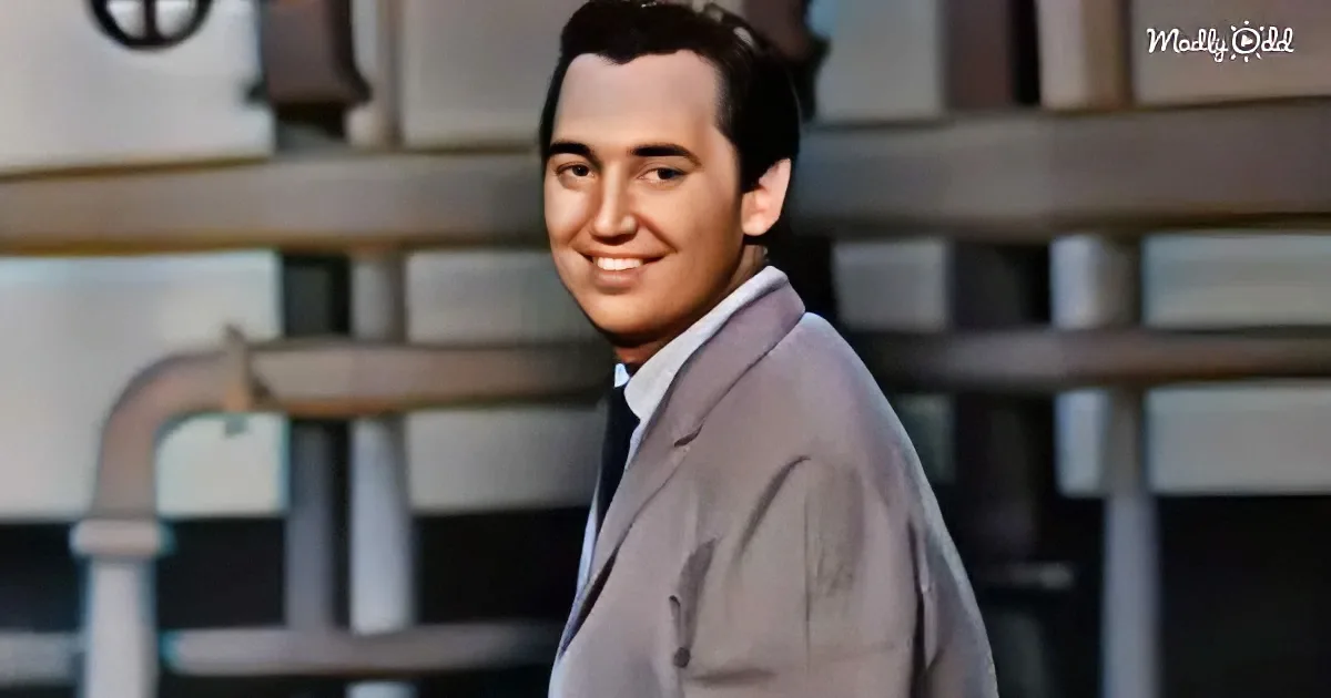 A man in a light gray suit and tie smiles, standing in front of a blurred background with shelves.