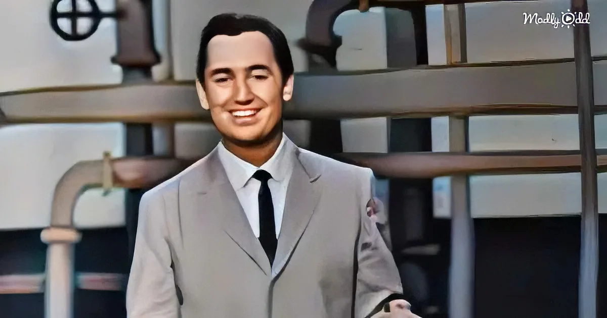 A man in a light gray suit and black tie smiles in front of a backdrop with pipes and machinery.