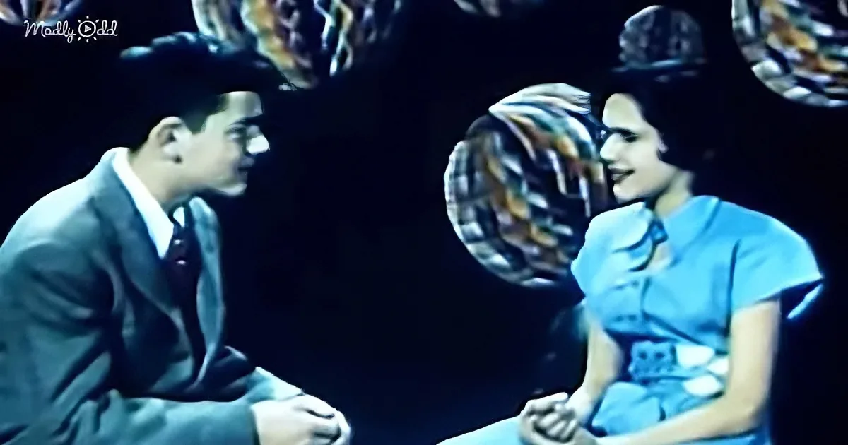 A man in a suit and a woman in a blue dress sit facing each other, surrounded by colorful, abstract patterns.