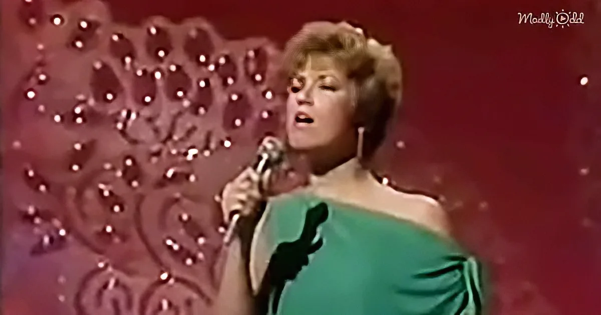 A woman in a green dress sings into a microphone on stage with a red, patterned backdrop.