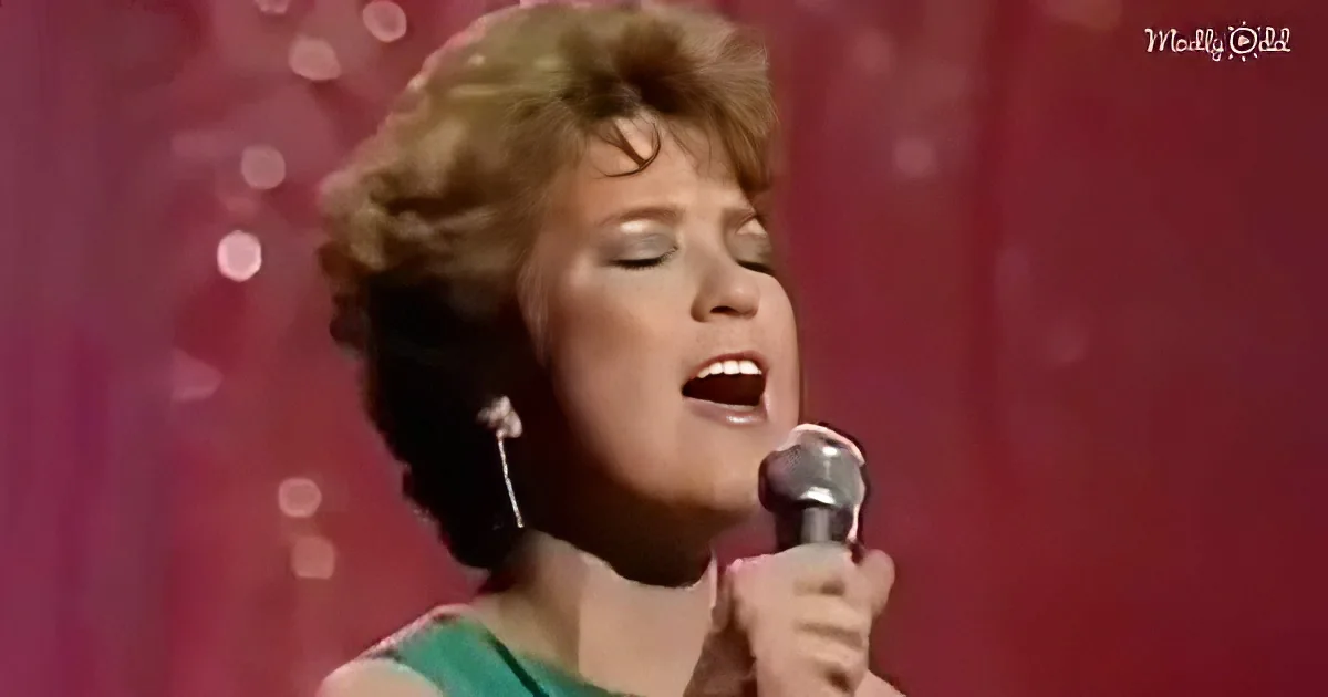 A woman with short, curly hair sings into a microphone against a red, sparkly background.