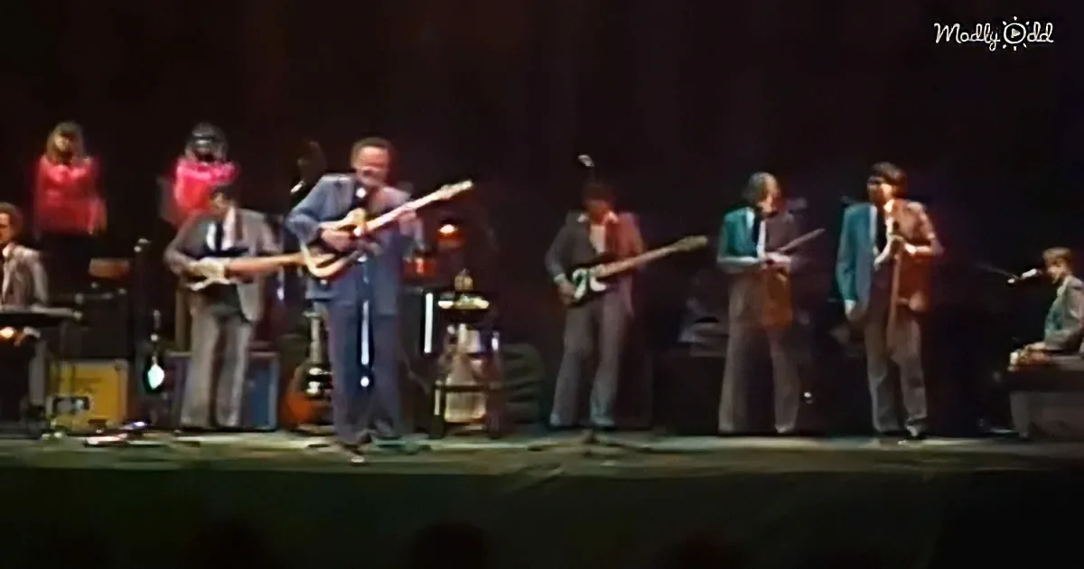A man in a suit plays guitar on stage with a band, surrounded by other musicians.