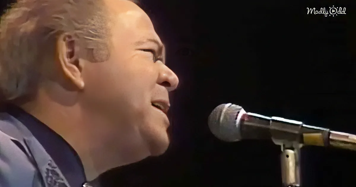 A man with short, light brown hair sings into a microphone, his face partially illuminated.