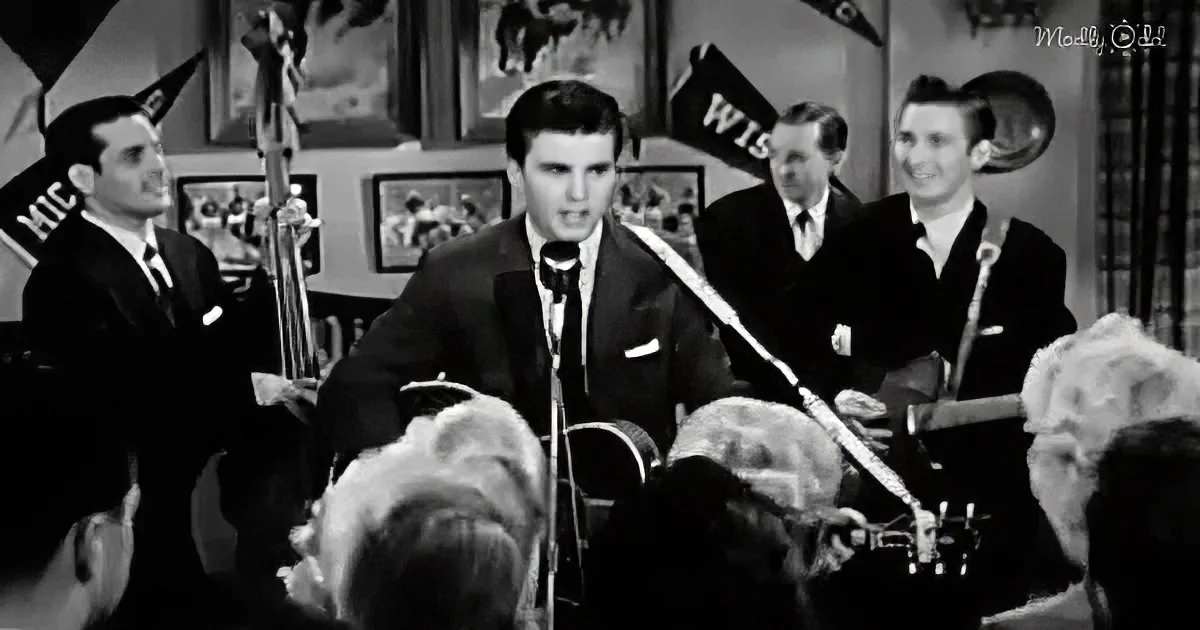 Elvis Presley performs on stage with a band, surrounded by a crowd and vintage decor.