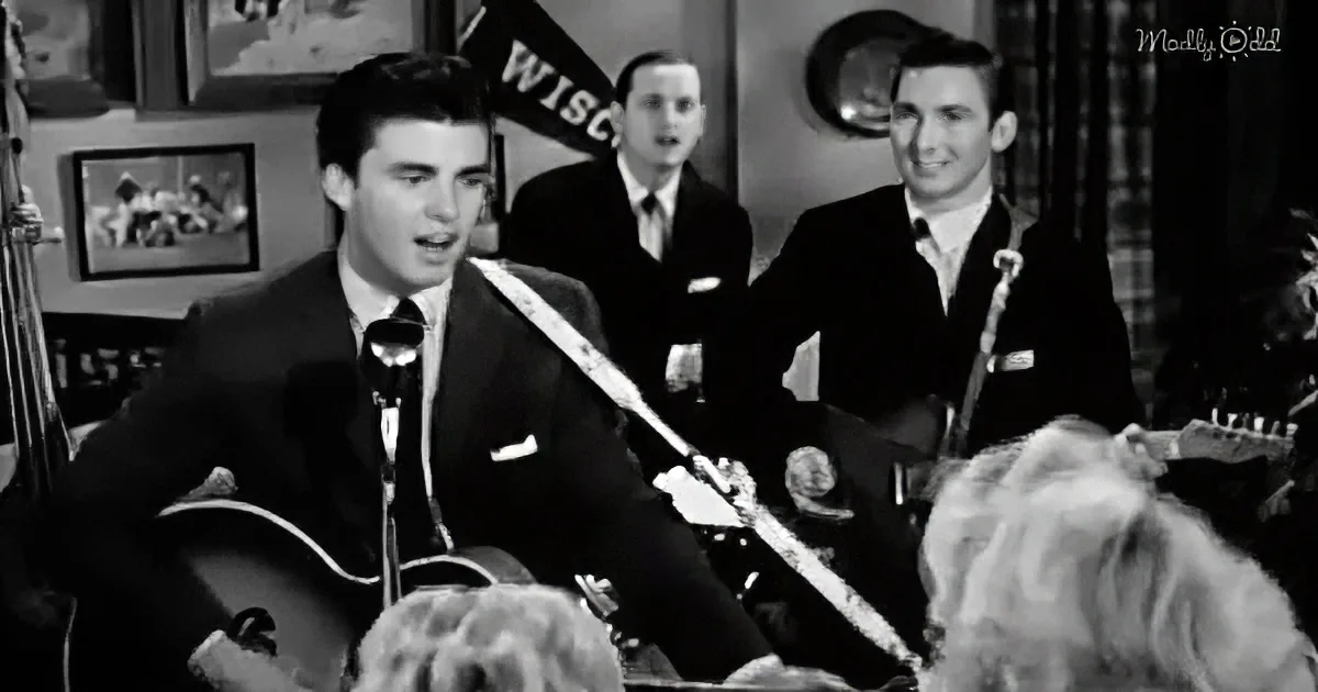 A man in a suit sings into a microphone, accompanied by a band, with a "WISC" flag in the background.