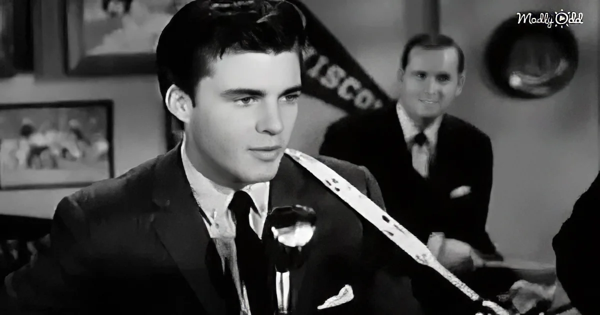 A man in a suit and tie speaks into a microphone, with another man in the background.
