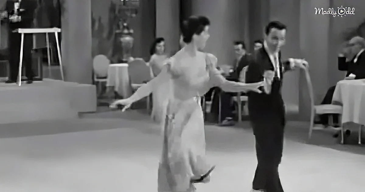 A couple in formal attire dances gracefully on a dance floor, surrounded by elegantly dressed onlookers.