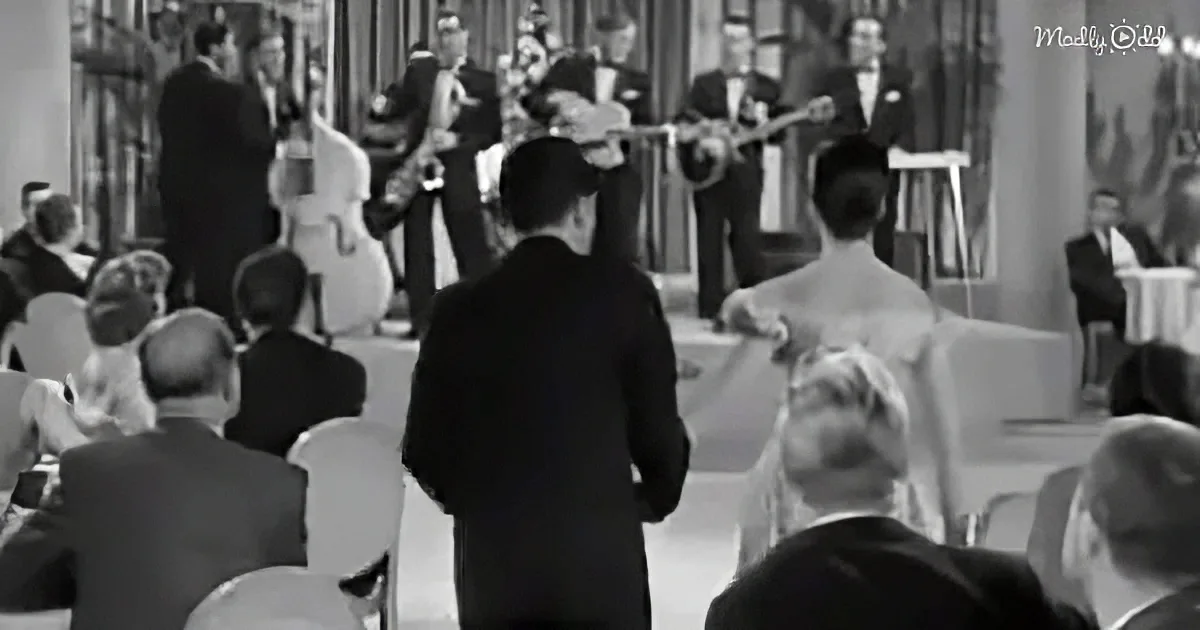 A man in a suit stands in front of a seated audience, facing a band playing in a formal setting.