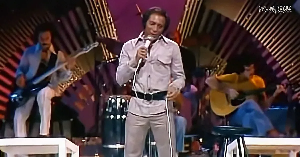 A man in a light-colored shirt and pants sings into a microphone on stage, accompanied by musicians.