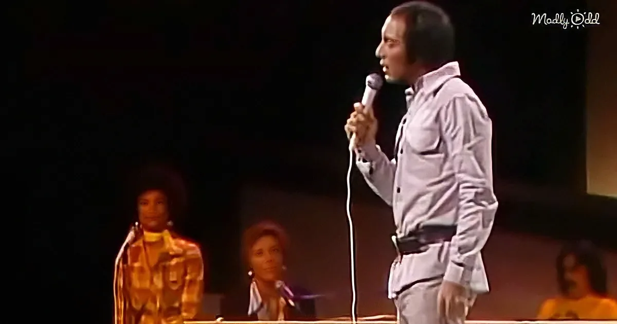 A man in a white shirt holds a microphone on stage, with a woman in a patterned dress standing nearby.
