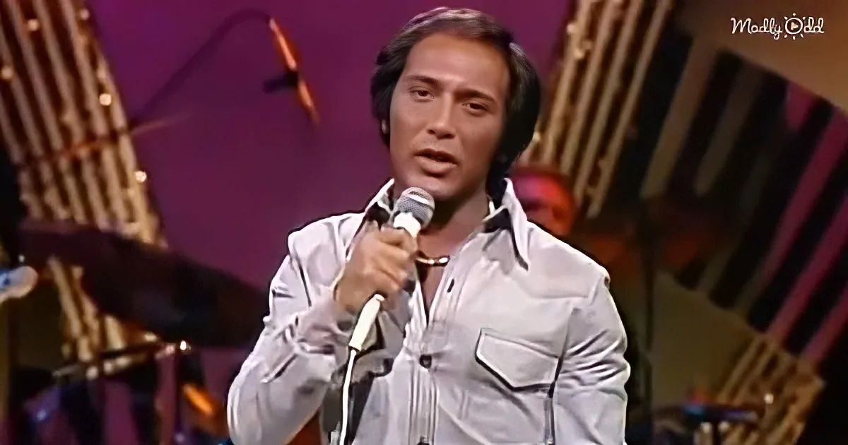 A man in a white shirt sings into a microphone on stage with a purple backdrop and musical instruments.