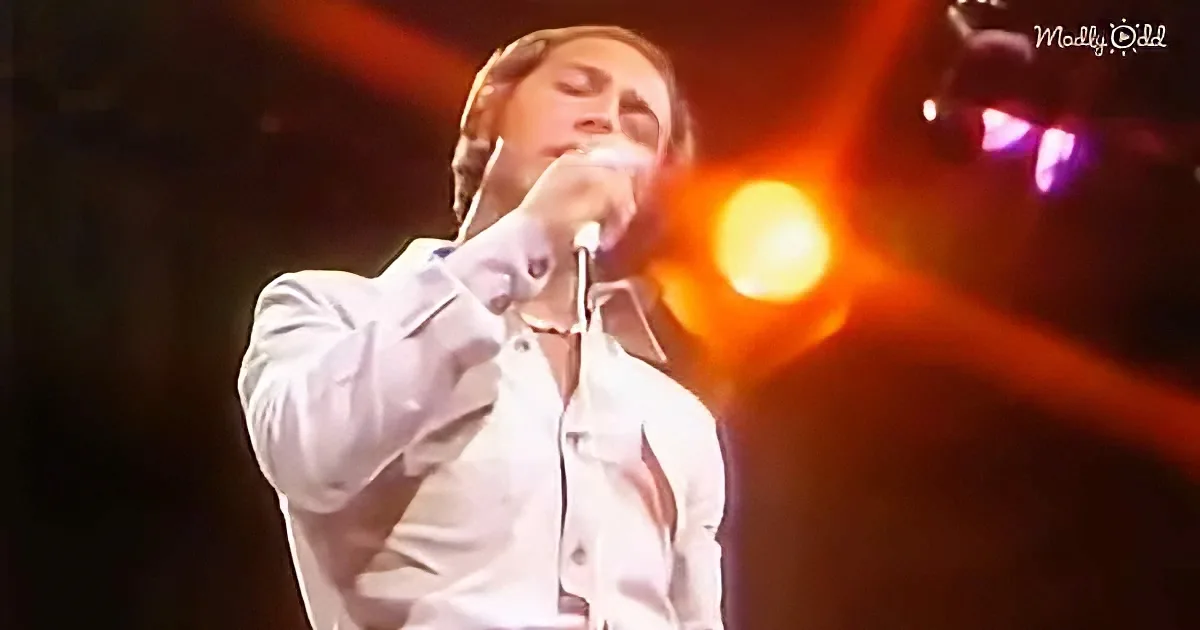 A person in a white shirt sings into a microphone on stage with bright stage lights.