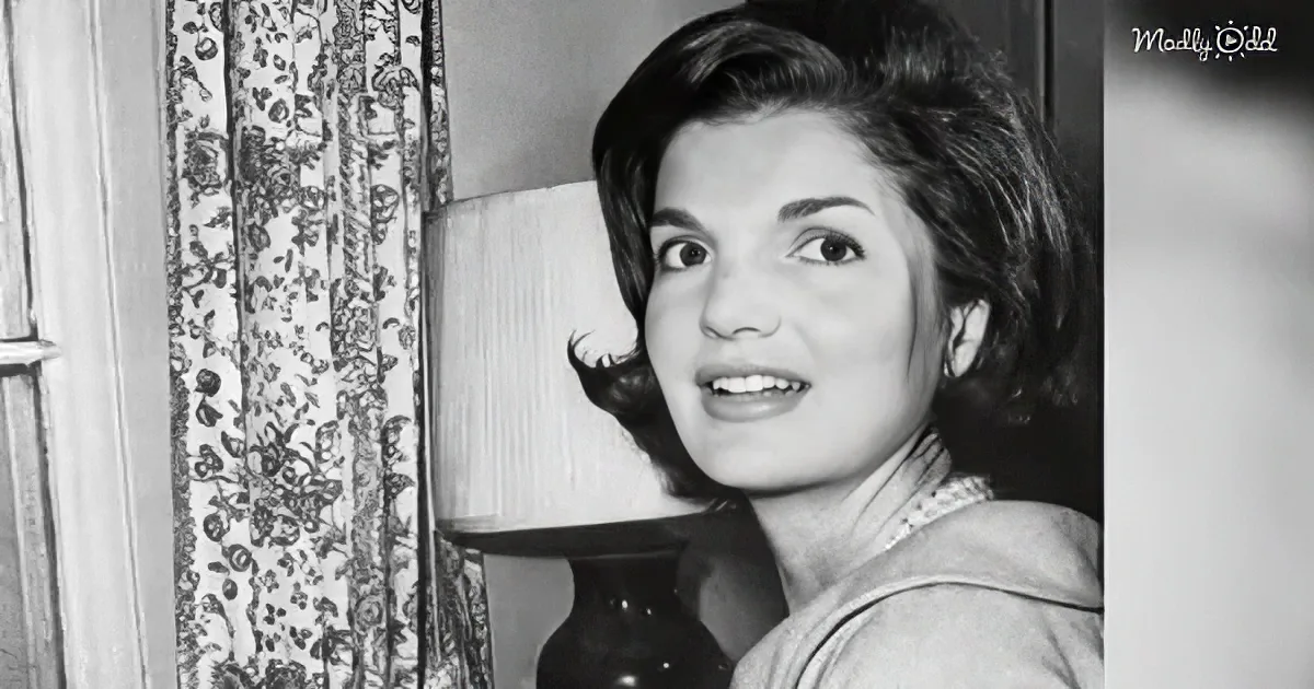 A woman with dark, styled hair smiles near a floral curtain and lamp.