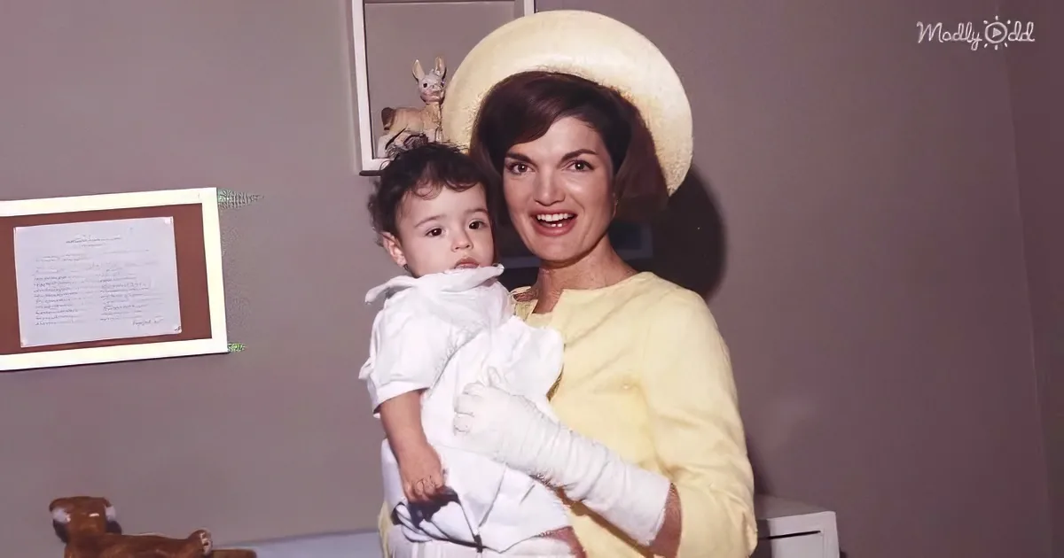 A woman in a yellow top holds a baby in white, both smiling, with a framed picture and stuffed animals in the background.