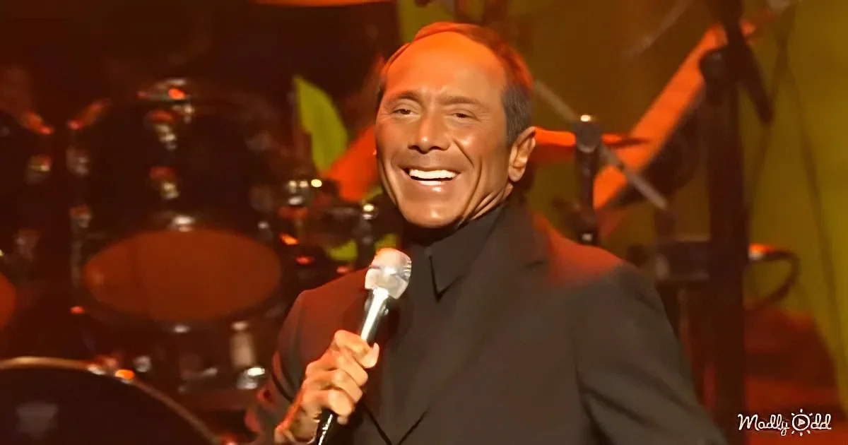 A man in a black suit smiles while holding a microphone on stage.