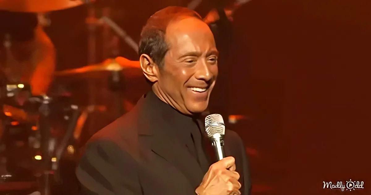 A man in a suit holds a microphone, smiling, with a band in the background.