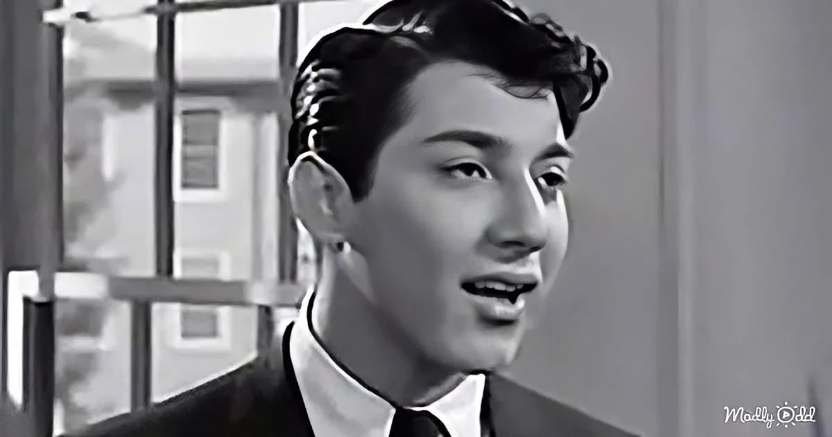 A man in a suit and tie stands in front of a window with bars, looking to the side.