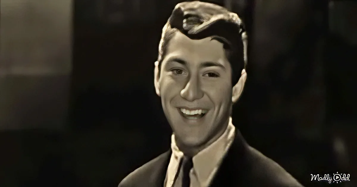 A smiling man in a suit and tie, looking to the side, with a dark background.