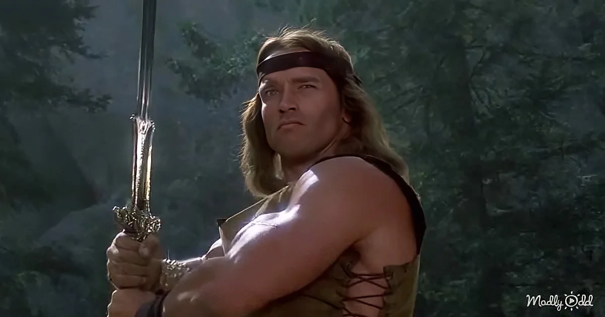 Arnold Schwarzenegger as Conan the Barbarian, holding a sword, wearing a headband and leather armor, standing in a forest.