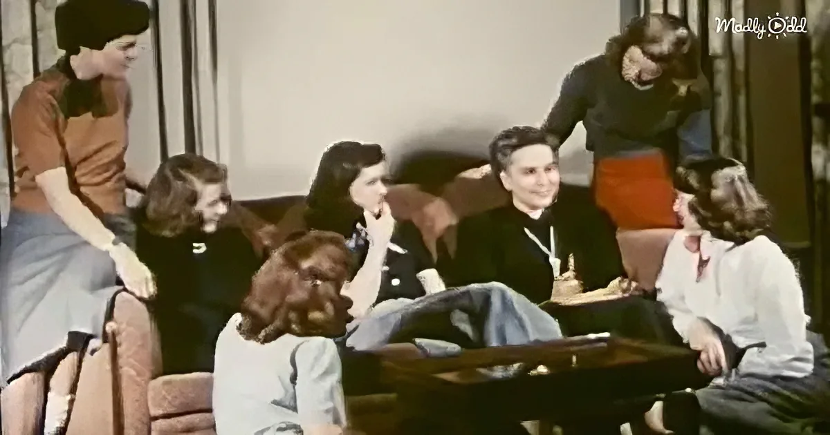 A woman in a black dress stands, leaning on a table, while others sit and converse in a vintage living room setting.