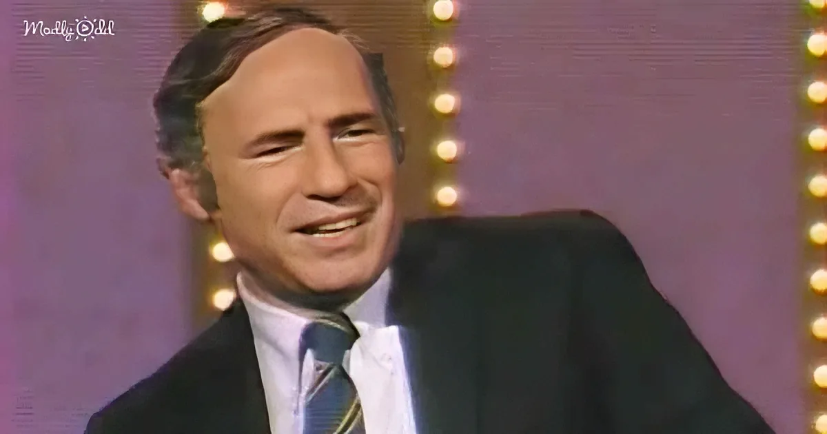A man in a suit and tie smiles, with a purple background and lights behind him.