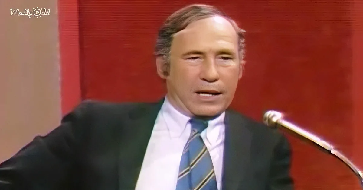 A man in a suit and tie speaks into a microphone against a red backdrop.