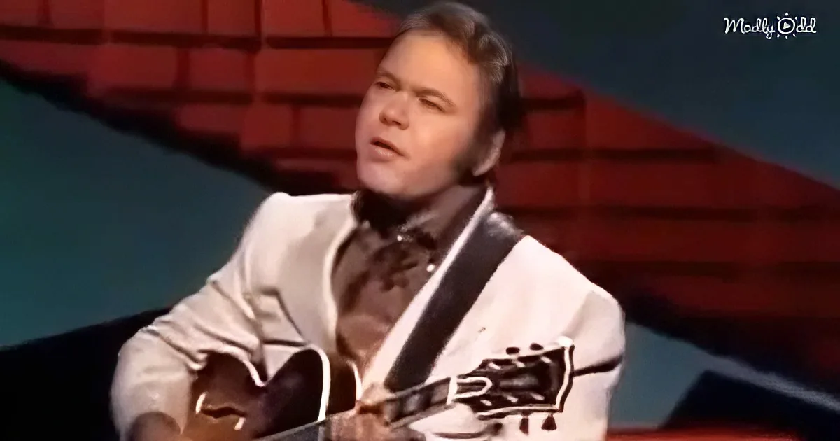 A man in a white suit plays a guitar on stage with a red brick background.