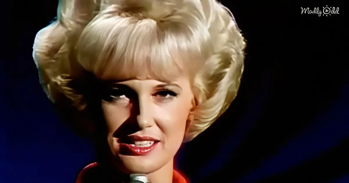 A blonde woman with a voluminous hairstyle and red lipstick is singing into a microphone.