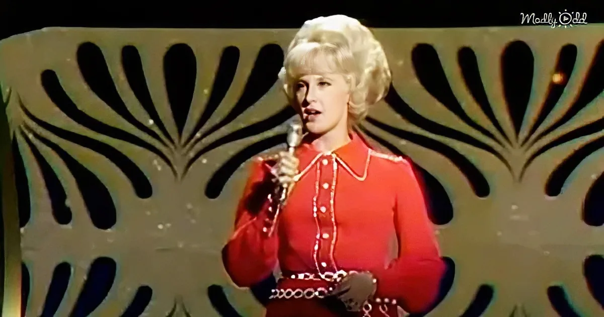 A person in a red outfit holds a microphone, standing in front of a decorative background.