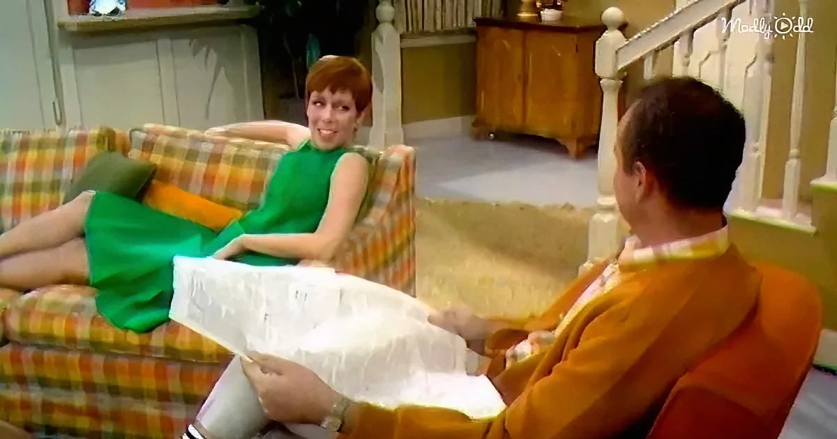 A woman in a green dress sits on a plaid couch, smiling at a man in an orange sweater holding a newspaper.
