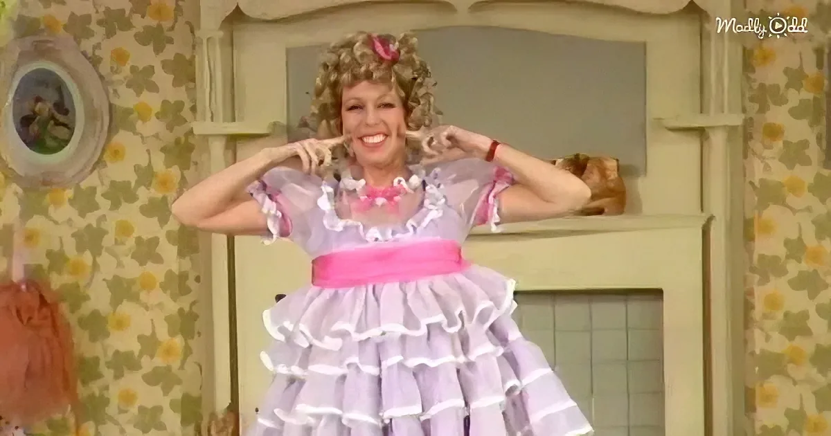 A woman in a purple ruffled dress with a pink sash smiles, holding her hair up, in a room with floral wallpaper and a mirror.