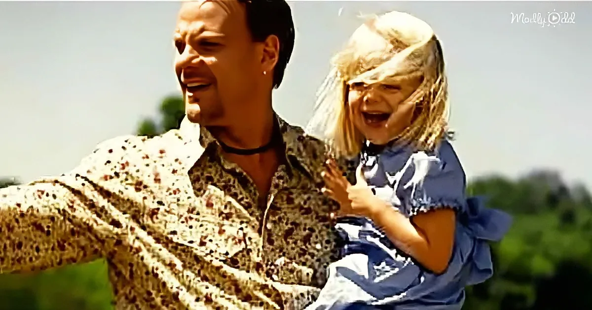A man in a patterned shirt holds a child in a blue dress, both laughing outdoors.