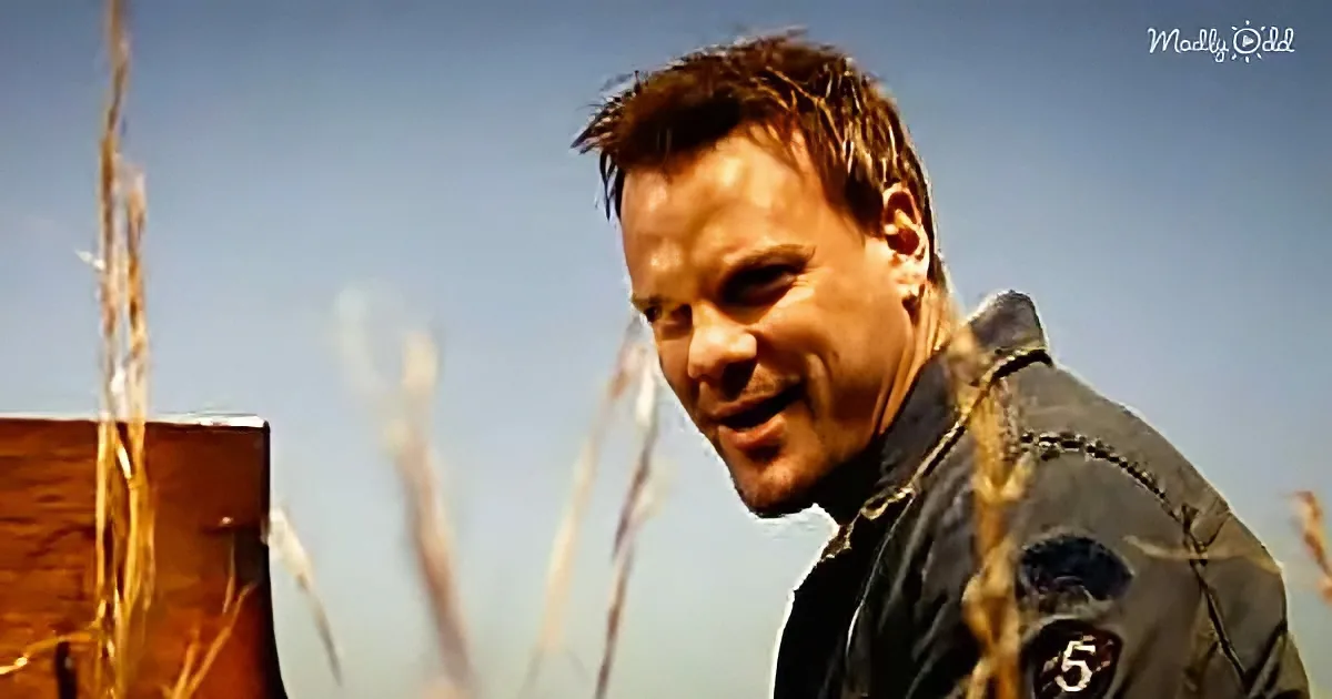 A man with short brown hair, wearing a dark jacket, smiles while standing in a field with tall grass and a clear sky.