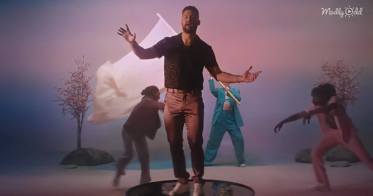 A man in a black shirt and brown pants stands with arms outstretched, surrounded by dancers in a foggy, surreal setting.