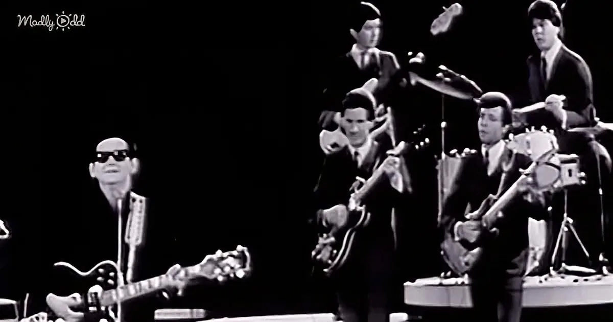 A man in sunglasses plays guitar on stage with a band, including a drummer and a guitarist.