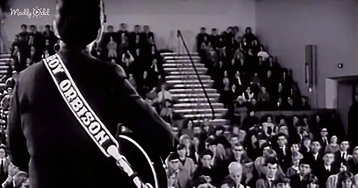 Roy Orbison, wearing a sash, stands on stage with a microphone, facing a crowd in a large auditorium.