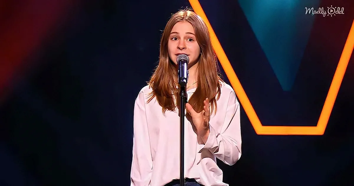 A person in a white shirt stands on stage with a microphone, gesturing with their hand.