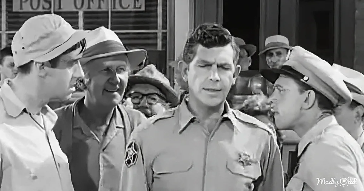 A man in a sheriff's uniform stands surrounded by others in a small-town setting, with a post office in the background.