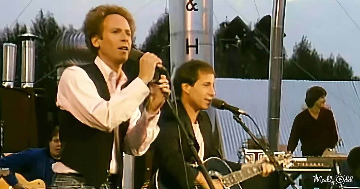 A man in a vest sings into a microphone while another plays guitar, with a band and audience in the background.