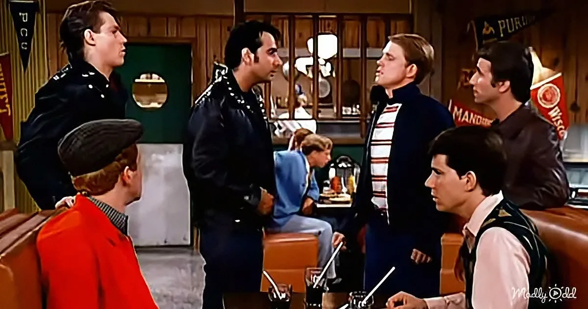 A man in a black leather jacket stands in a room with others, possibly in a bar or lounge setting.