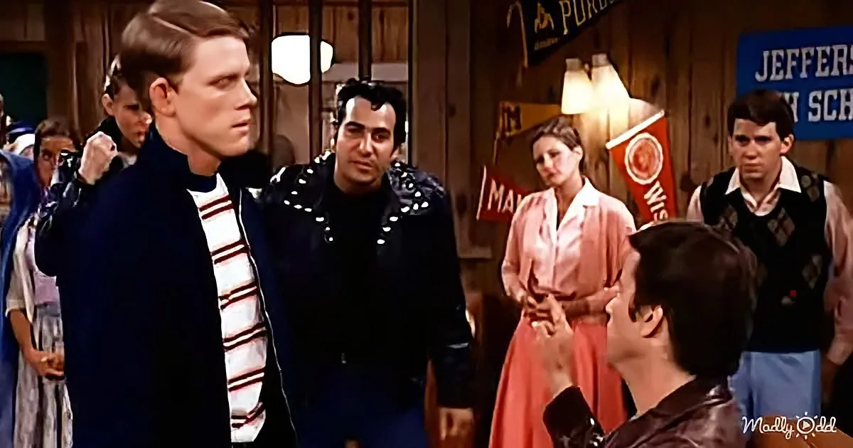 A man in a striped shirt stands facing another man in a leather jacket, surrounded by onlookers in a school setting.