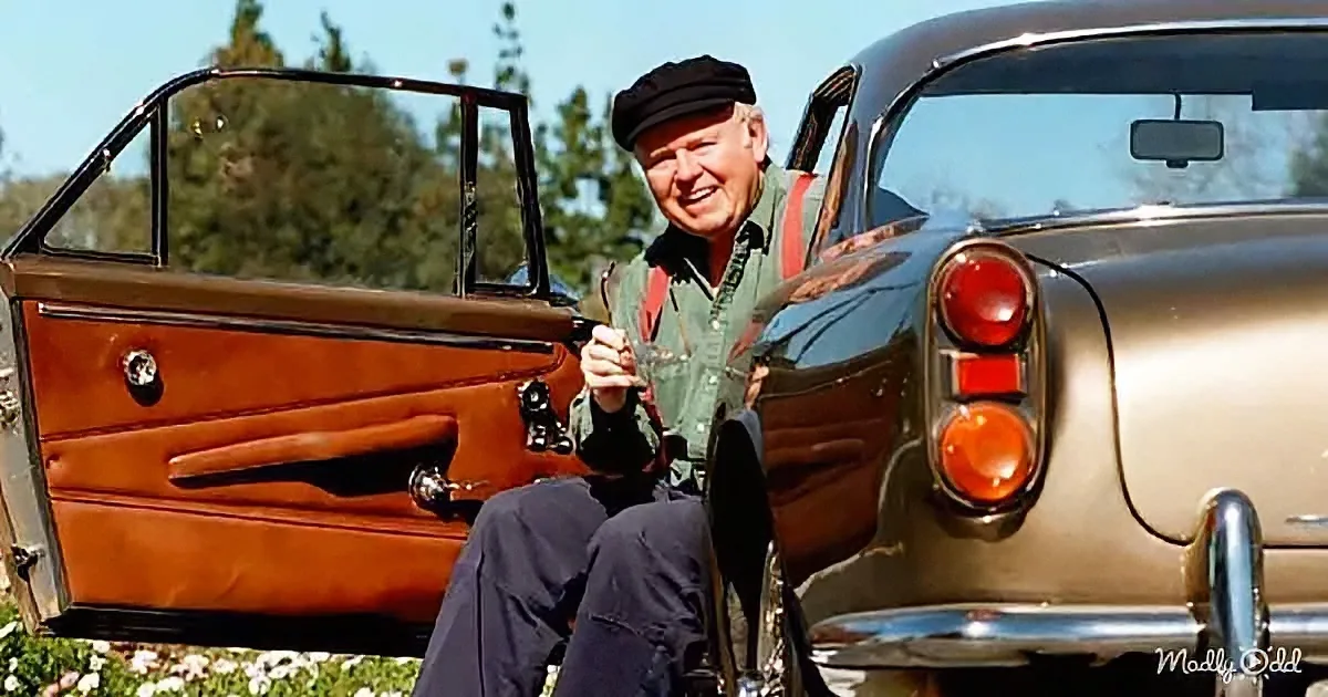 A man in a green shirt and black cap sits beside a vintage car with an open door, holding a drink.