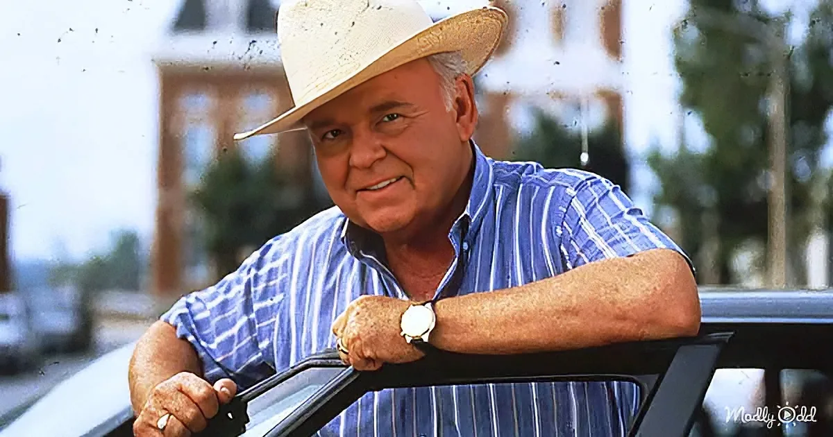 A man in a straw hat and striped shirt leans on a car, smiling, with a blurred background of trees and buildings.