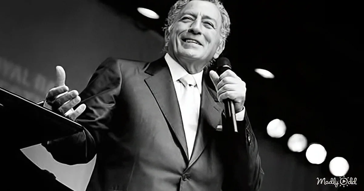 A man in a suit holds a microphone, giving a thumbs-up, with a dark background and stage lights.