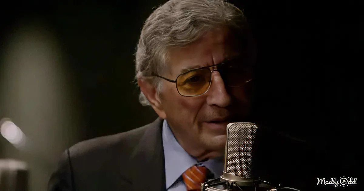A man in a suit and tie sings into a microphone, wearing glasses, in a dimly lit setting.