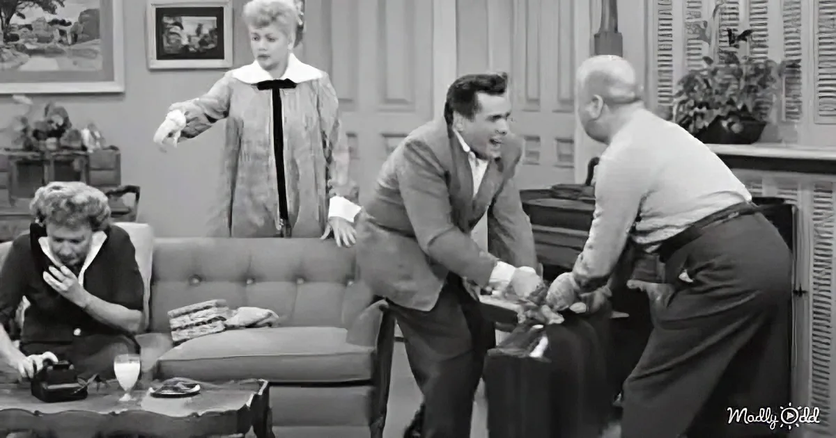 A man in a suit shakes hands with another, while a woman in a dress and another woman sit in the background.
