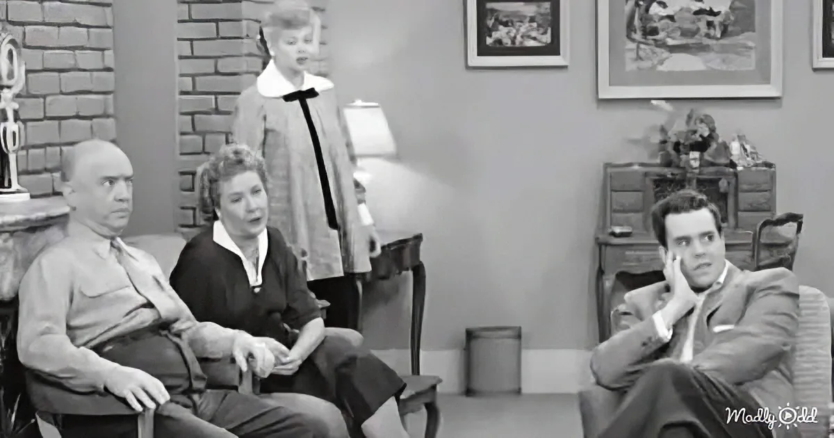 A man in a suit sits in a chair, hand on face, while others watch in a living room setting.