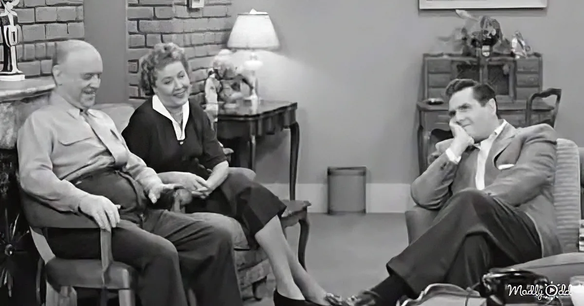 Three people in a living room, two men and a woman, seated and smiling, with a lamp and furniture in the background.