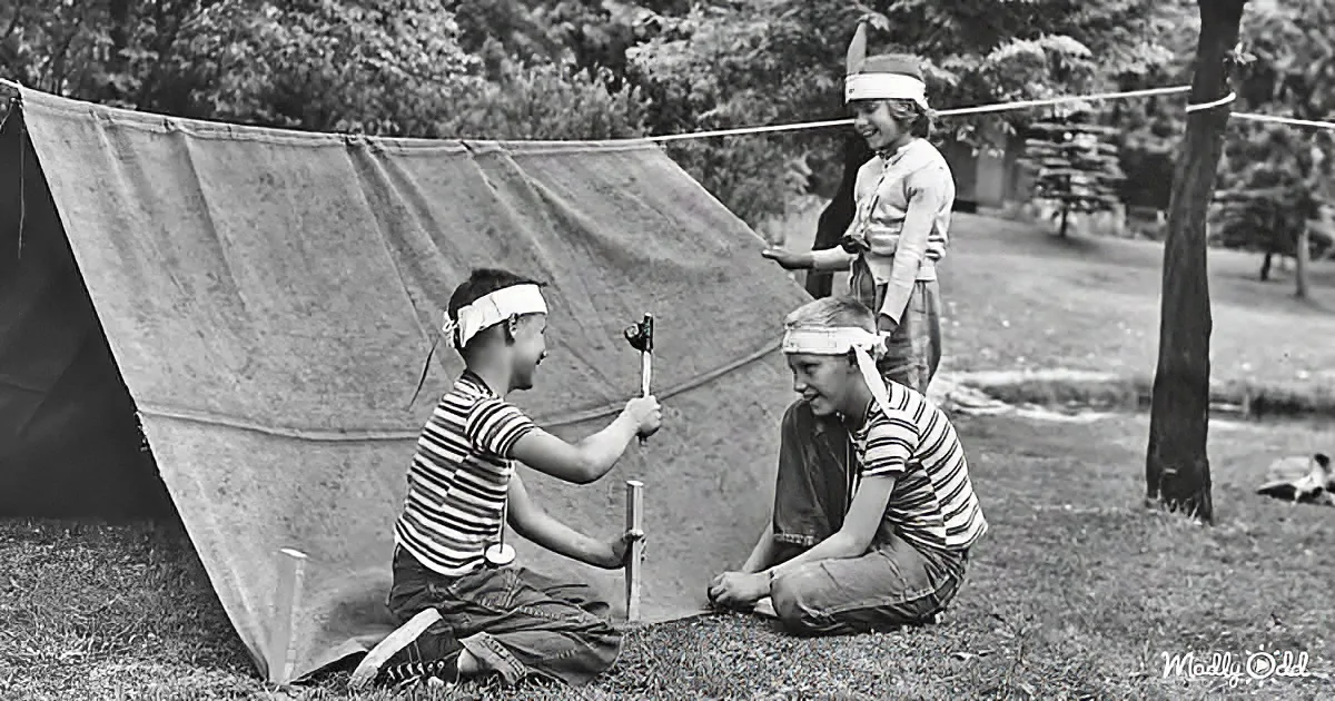 Three children in striped shirts and headbands play with sticks near a tent in a grassy area.